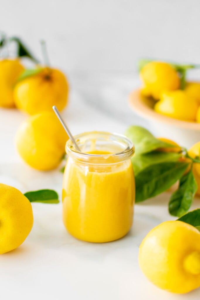 homemade organic lemon curd with egg yolks in a glass jar on a marble counter with fresh lemons.