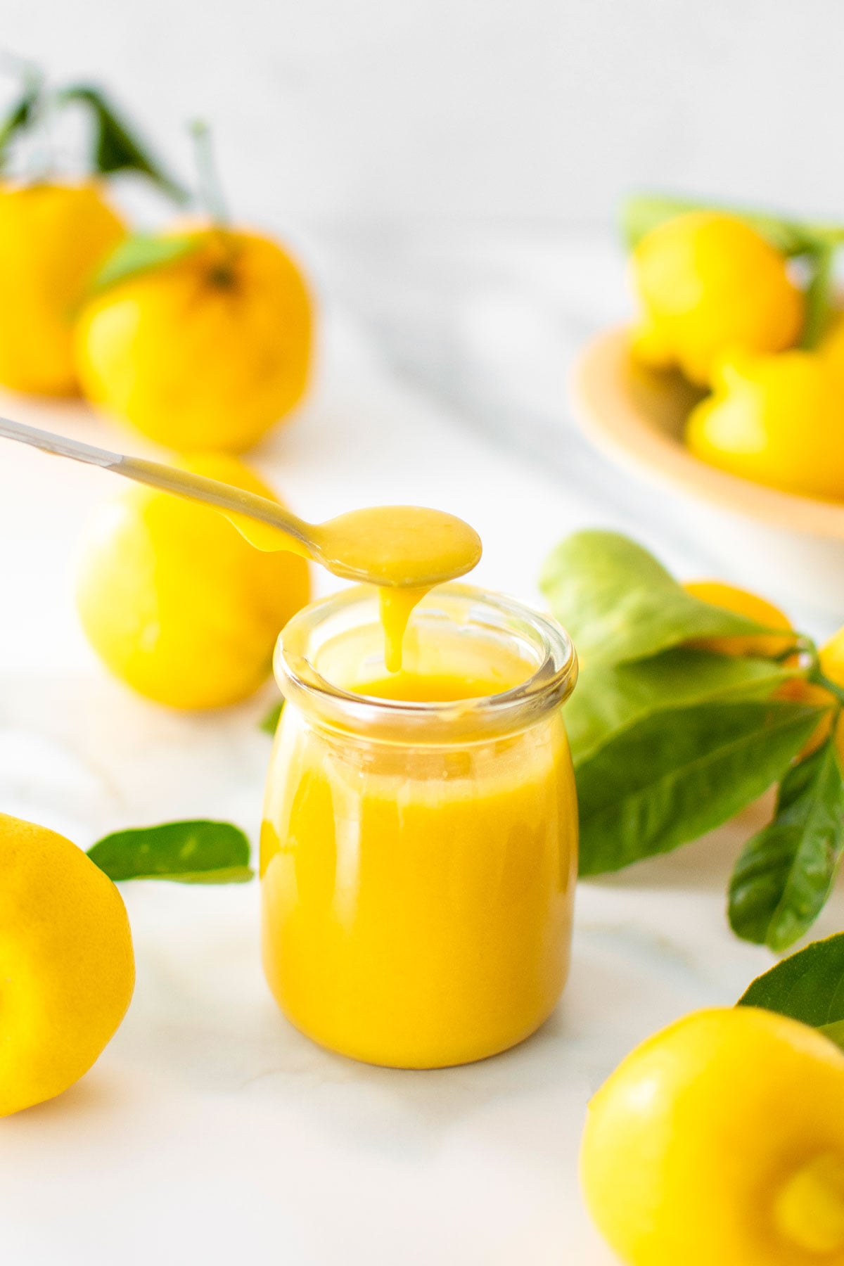 homemade organic lemon curd in a small glass jar with a spoon lifting some out on a marble counter with lemons.