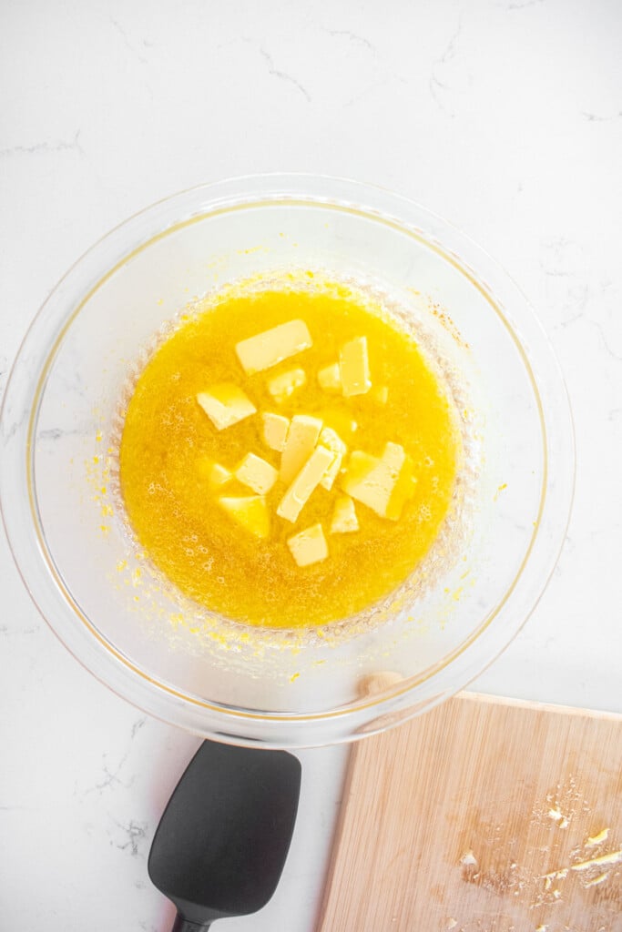 butter cubes being added into lemon curd in a glass mixing bowl on a white marble counter.