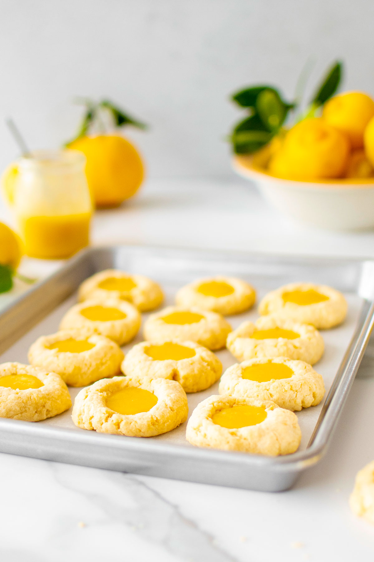 baking sheet with lemon curd coconut thumbprint cookies on a marble counter with fresh lemons and lemon curd.