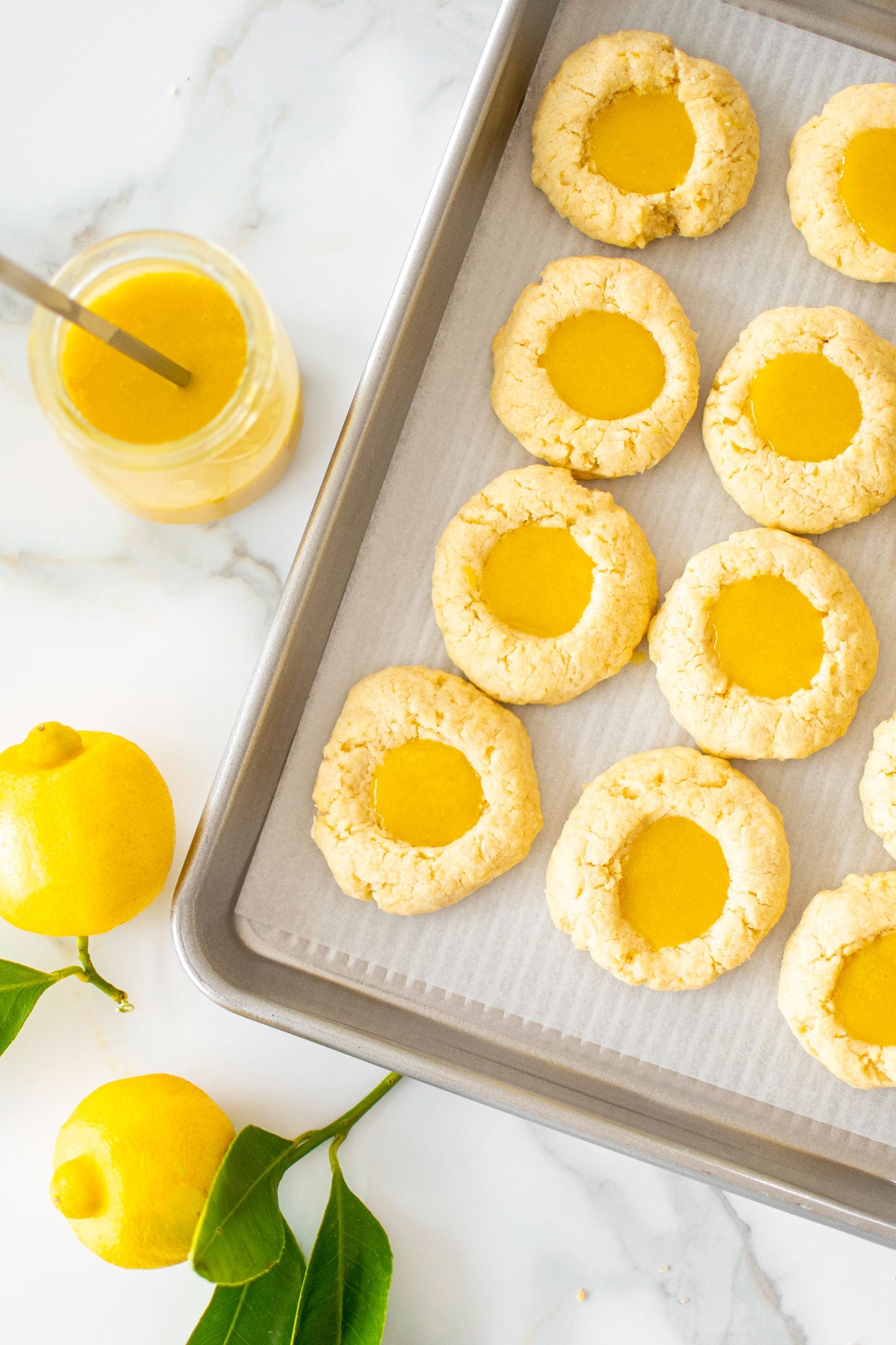baking sheet with lemon curd coconut thumbprint cookies on a marble counter with fresh lemons and lemon curd.