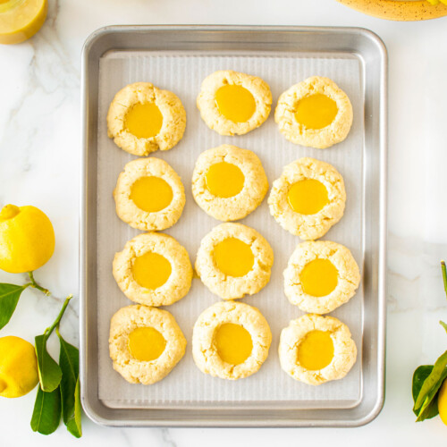 baking sheet with lemon curd coconut thumbprint cookies on a marble counter with fresh lemons and lemon curd.