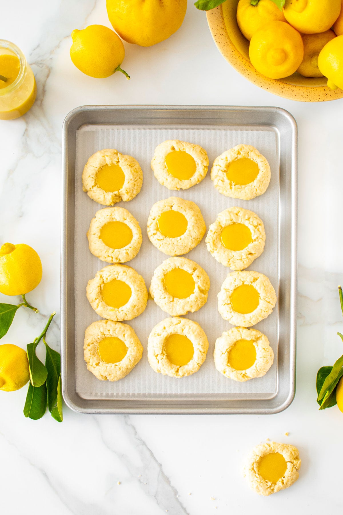 baking sheet with lemon curd coconut thumbprint cookies on a marble counter with fresh lemons and lemon curd.