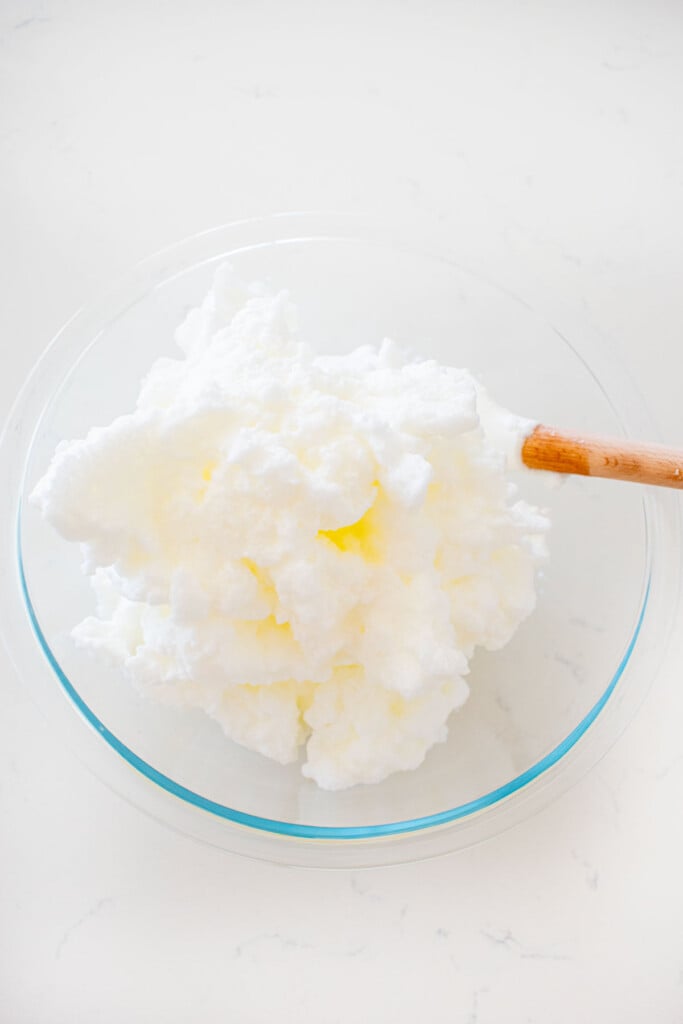 whipped egg whites to stiff peaks in a glass mixing bowl with a spatula on a white marble counter.