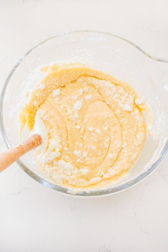 egg whites being folded into a white cake batter in a glass stand mixing bowl on a white marble counter.