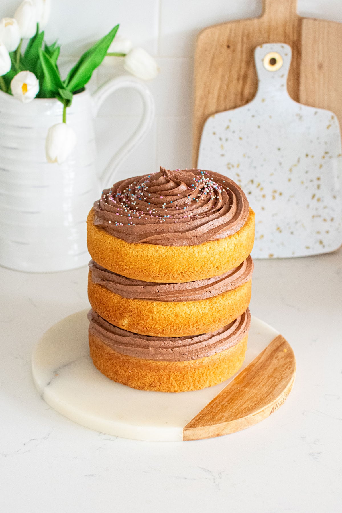 simple naked birthday cake with chocolate frosting and rainbow sprinkles on a marble cake plate on kitchen counter.