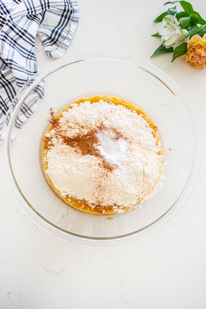 glass mixing bowl with dry ingredients added to wet ingredients for apple cinnamon cupcakes on a marble counter with fresh flowers and a kitchen towel.