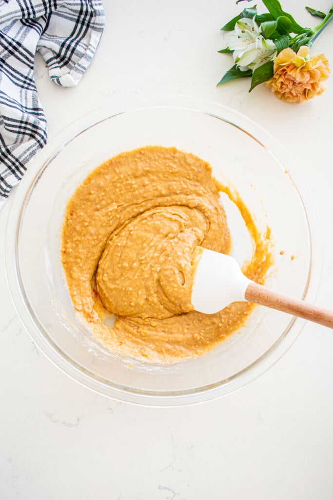 glass mixing bowl with apple cinnamon cupcake batter with a spatula in it on a marble counter with fresh flowers and a kitchen towel.