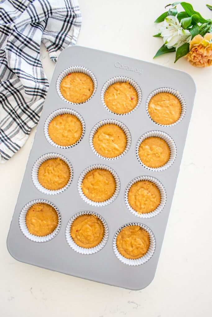 cupcake pan with liners filled with apple cinnamon cupcake batter on a marble counter with fresh flowers and a kitchen towel.
