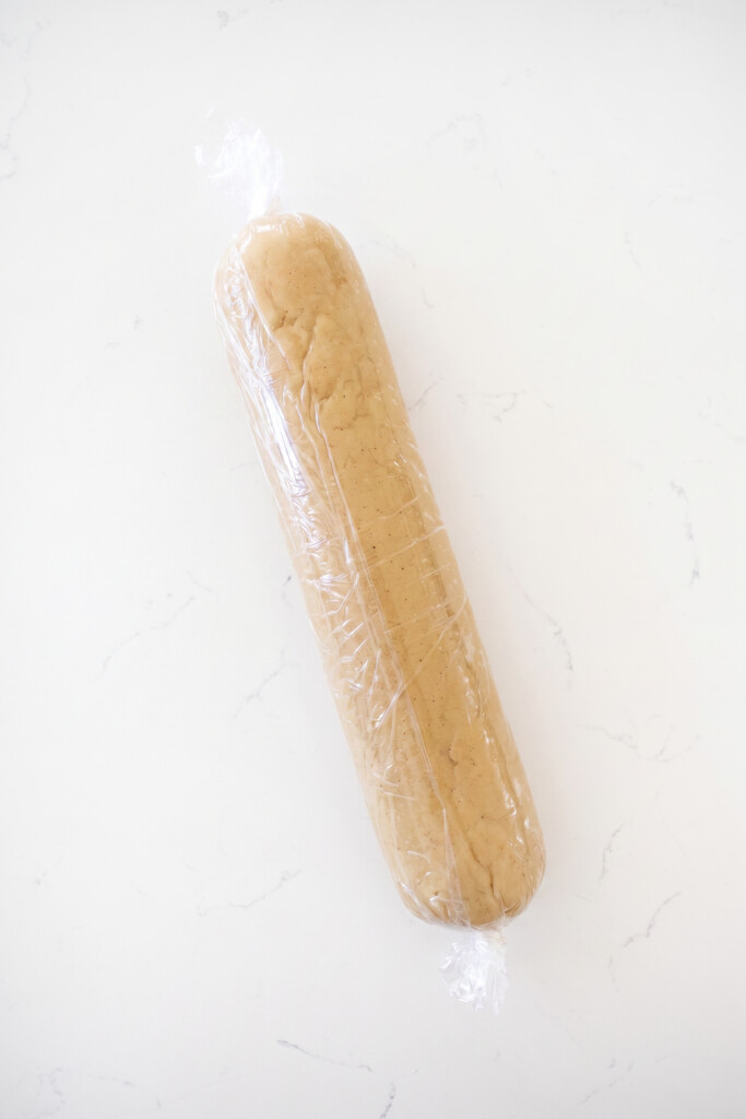 brown sugar shortbread cookie dough log wrapped in plastic wrap on a white marble counter.