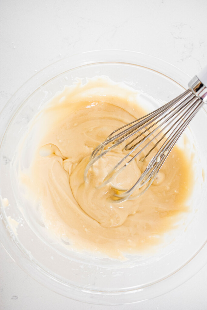 maple cream cheese icing in a glass mixing bowl with a whisk on a white marble counter.