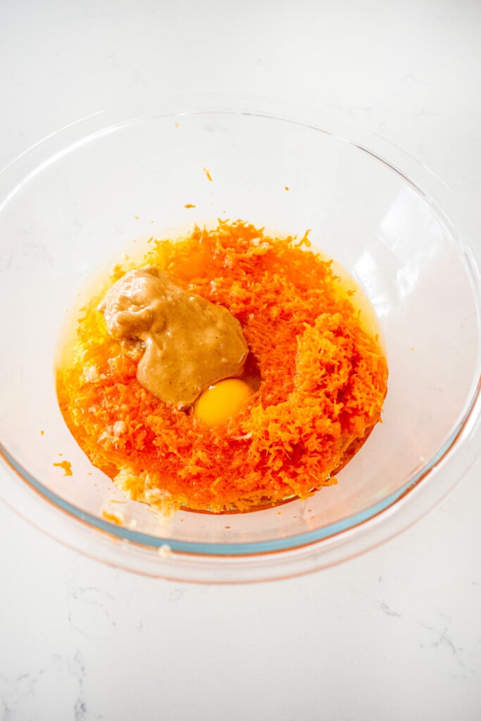 grated carrots, apples, peanut butter, egg and coconut oil in a glass mixing bowl on a white marble counter.