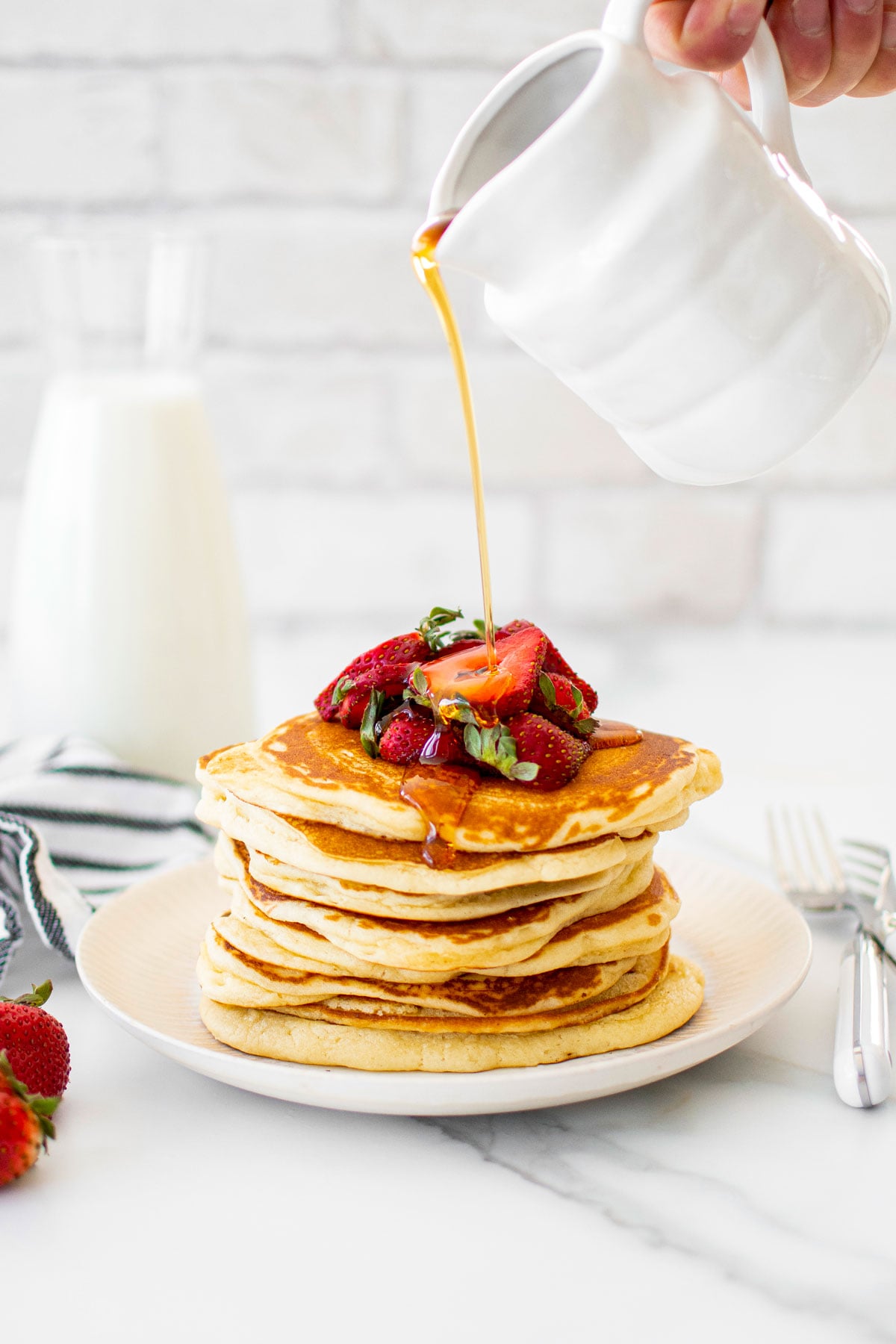 maple syrup being poured on a stack of homemade buttermilk pancakes on a plate with sliced strawberries on a white marble counter.