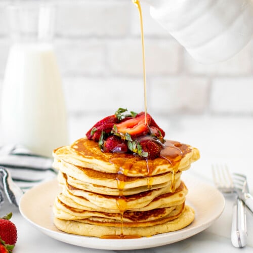 maple syrup being poured on a stack of homemade buttermilk pancakes on a plate with sliced strawberries on a white marble counter.