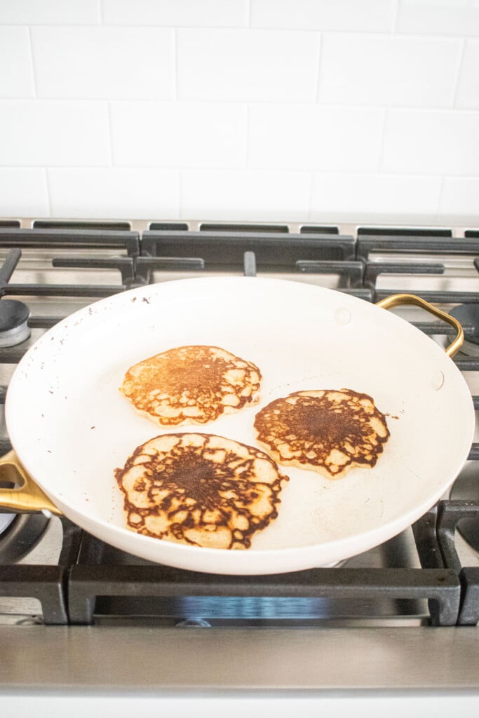 large white skillet on the stovetop cooking three buttermilk pancakes.