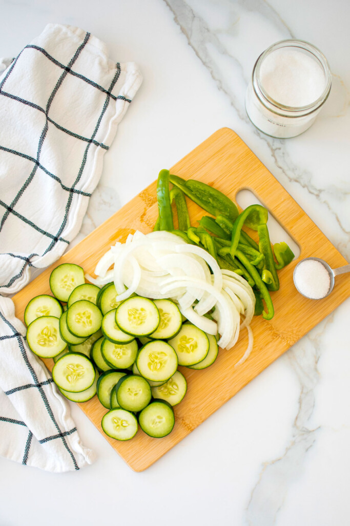 ingredients of refrigerator pickles sliced on a wood cutting board on a white marble countertop.