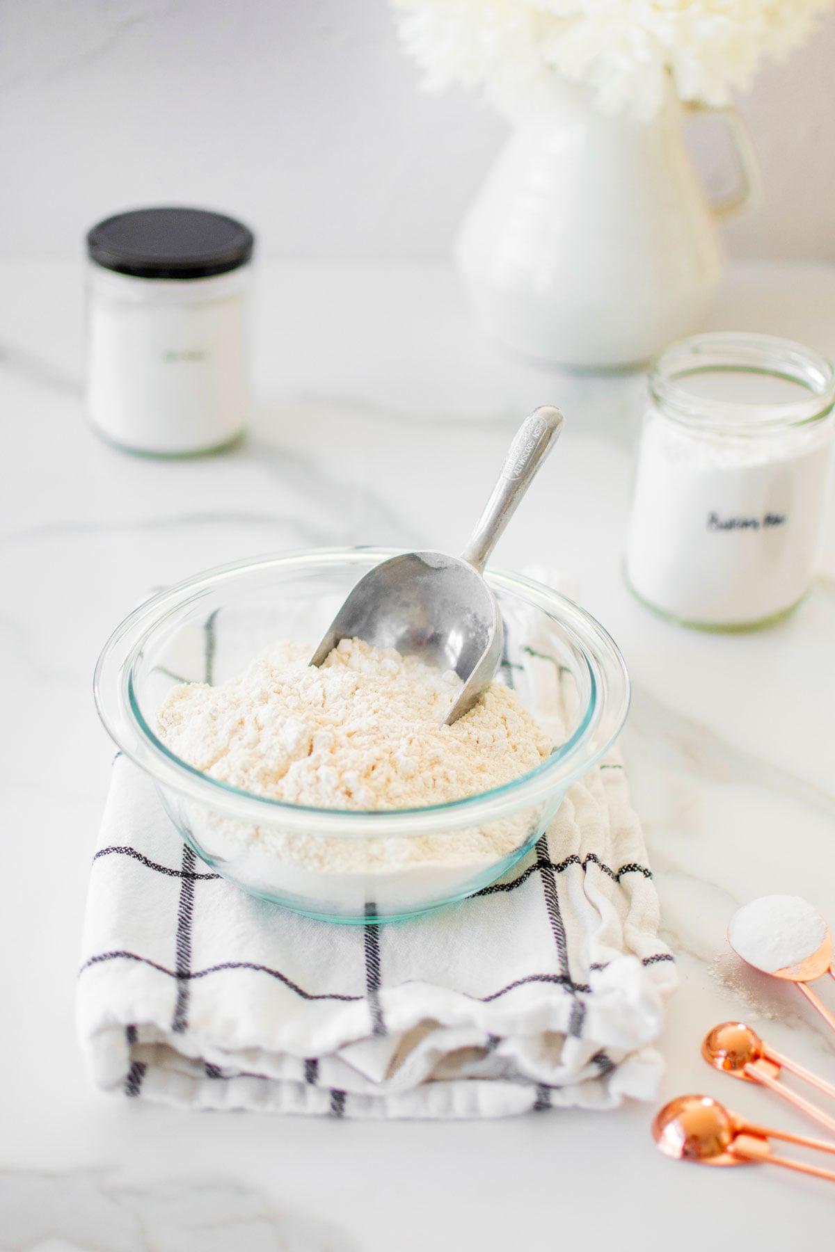 glass bowl of self rising flour on a kitchen towel on a marble table with a flour scoop in it.