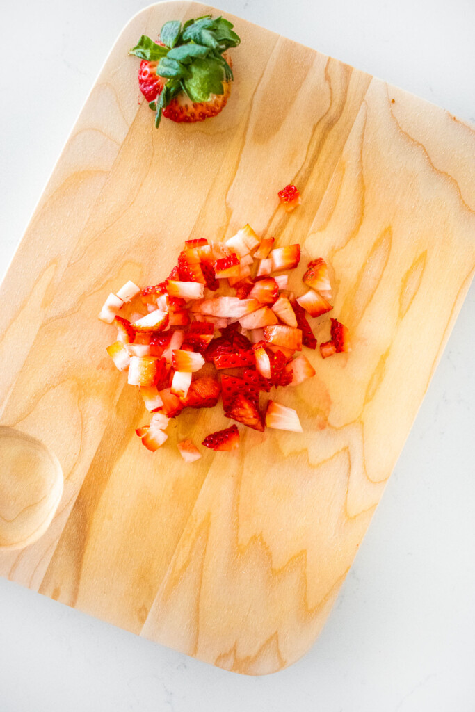 chopped strawberries on a wood cutting board on a white marble counter.
