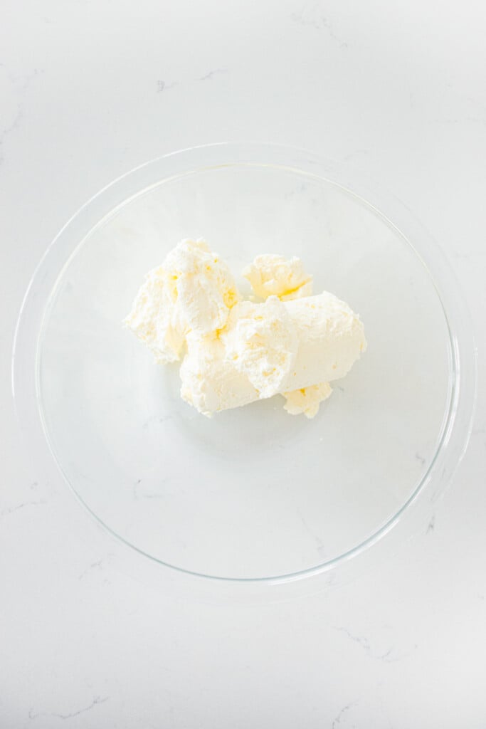 glass mixing bowl with cream cheese on a white marble counter.