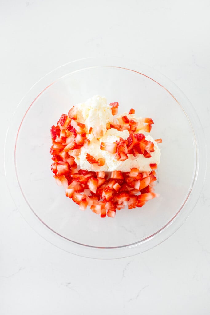 glass mixing bowl with cream cheese and chopped strawberries on a white marble counter.