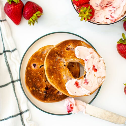 bagel with strawberry cream cheese spread on a white plate on a marble counter.