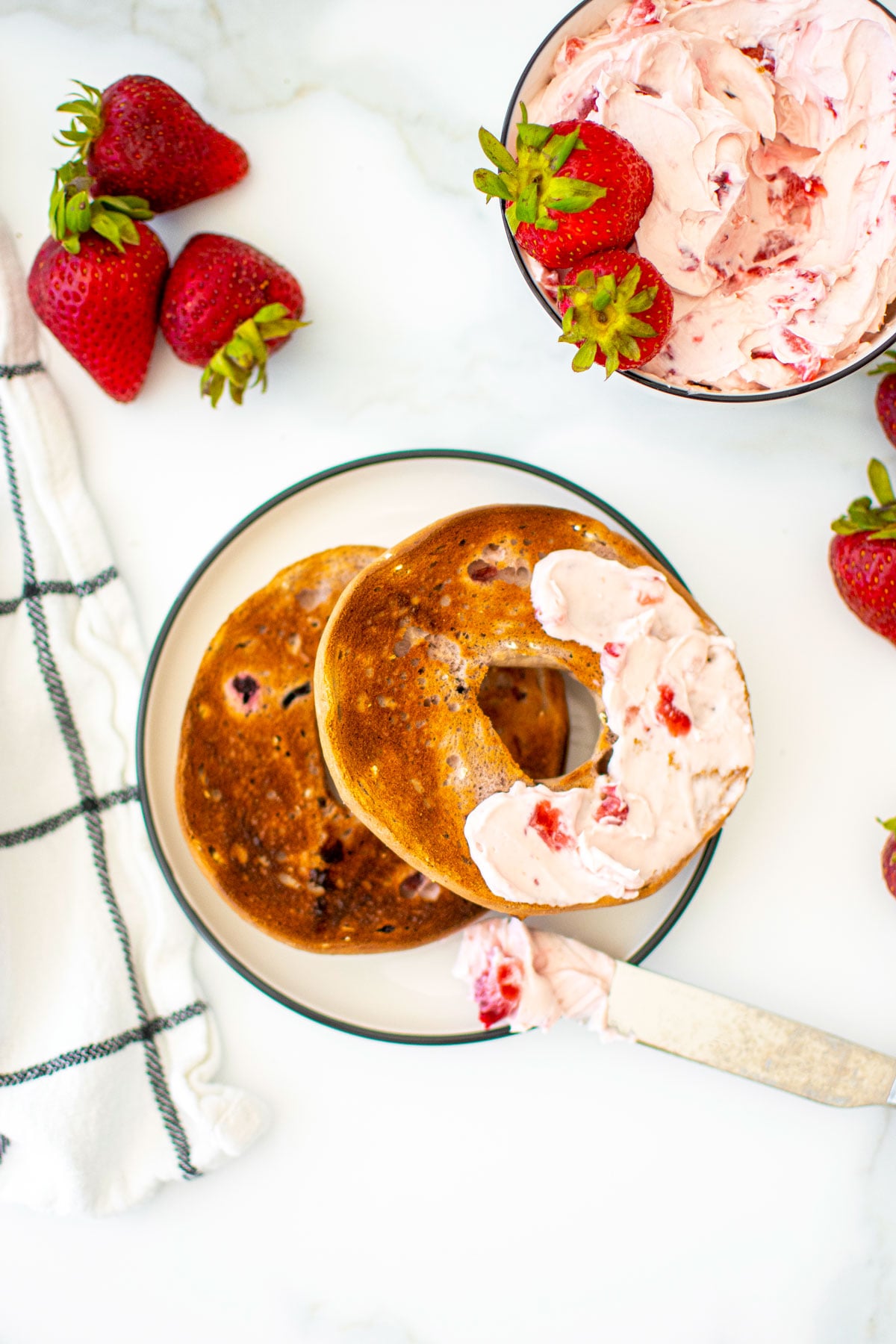bagel with strawberry cream cheese spread on a white plate on a marble counter.