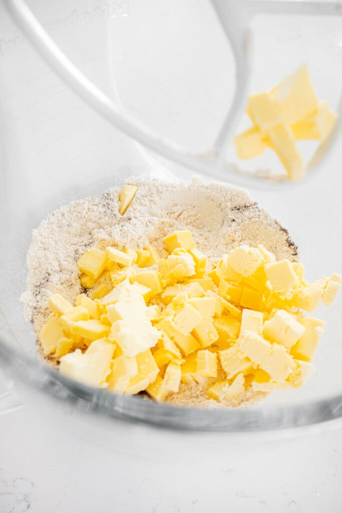 chopped cold butter on top of flour in a glass mixing bowl on a white marble counter.