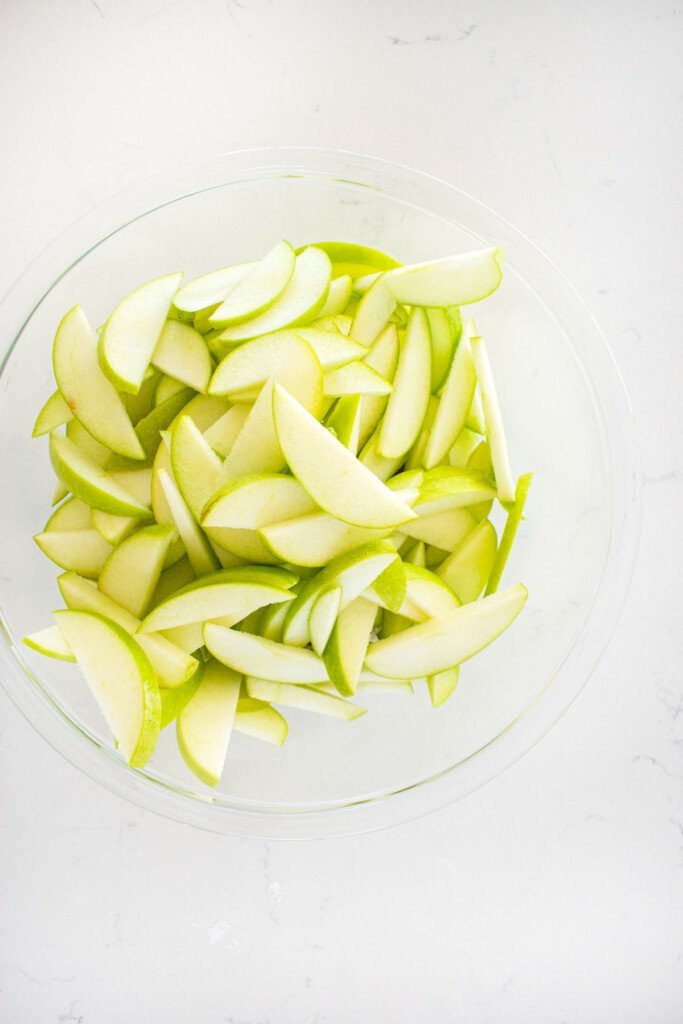sliced granny smith apples in a glass mixing bowl on a white marble counter.