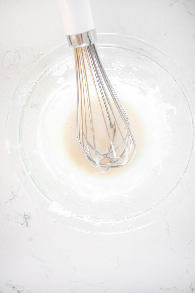 glaze mixed in a glass mixing bowl with a whisk on a white marble counter.