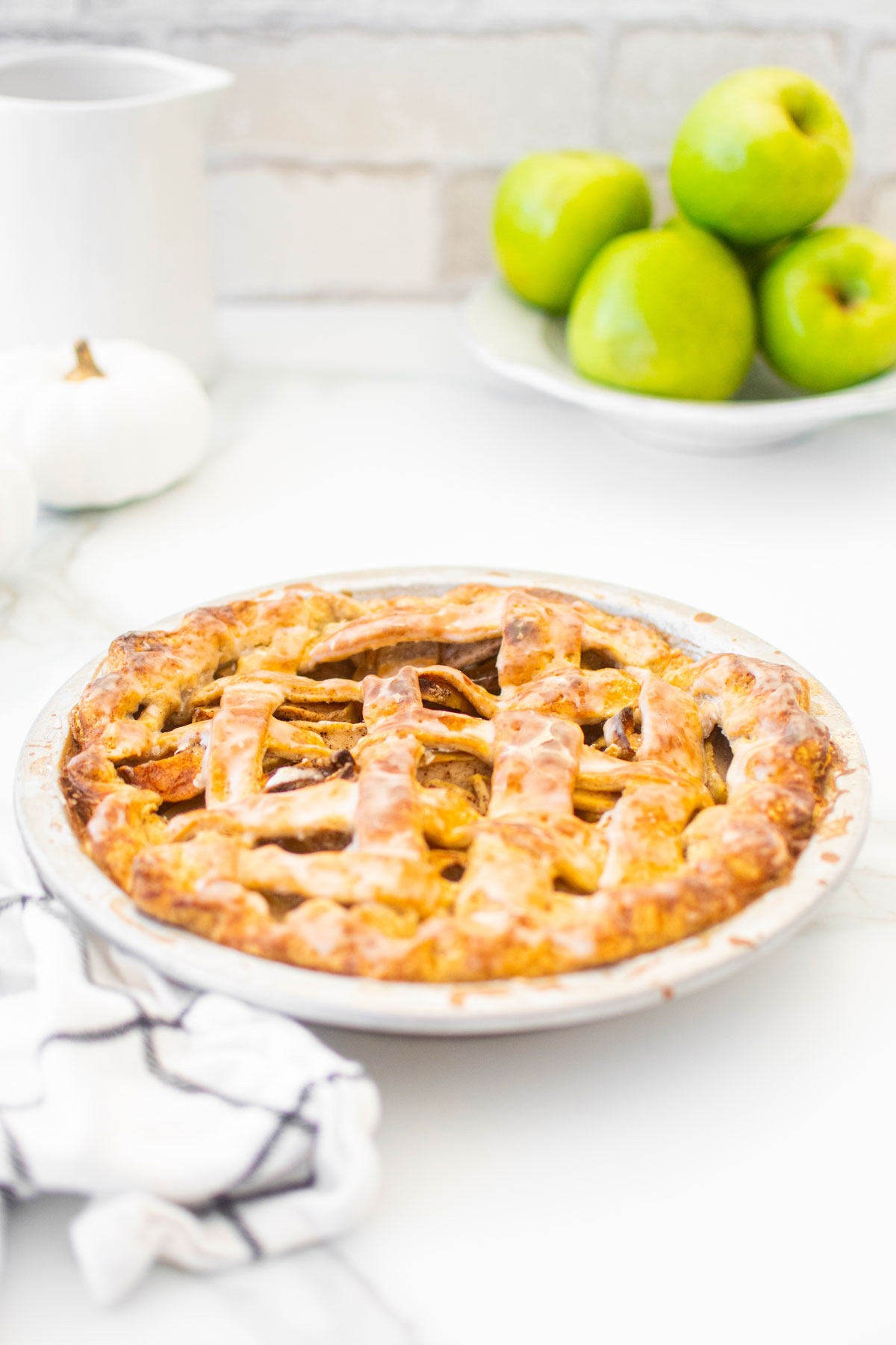 glazed vanilla bean apple pie on a white marble counter with green apples and white pumpkins.