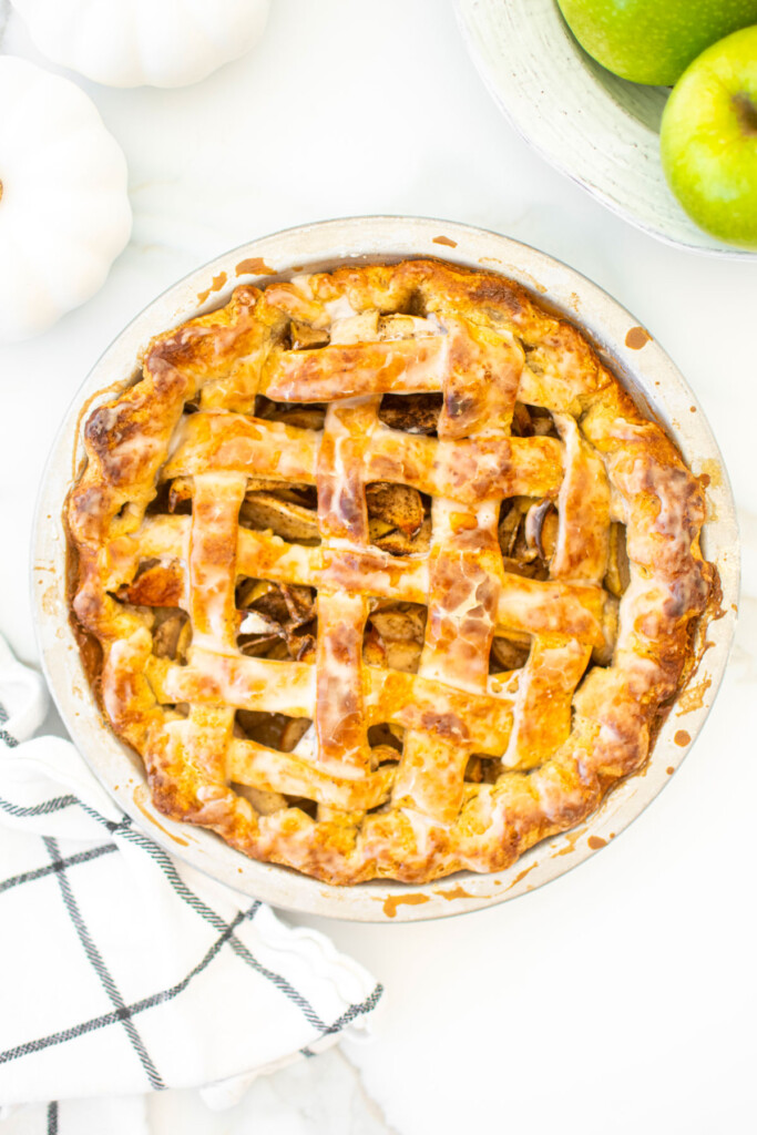 glazed vanilla bean apple pie on a white marble counter with green apples and white pumpkins.