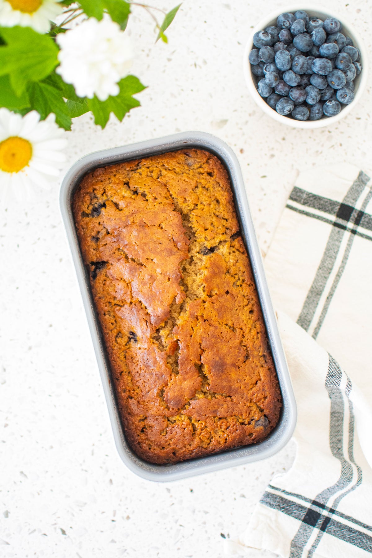 blueberry buttermilk banana bread in a loaf pan on a white stone countertop with fresh blueberries and flowers.