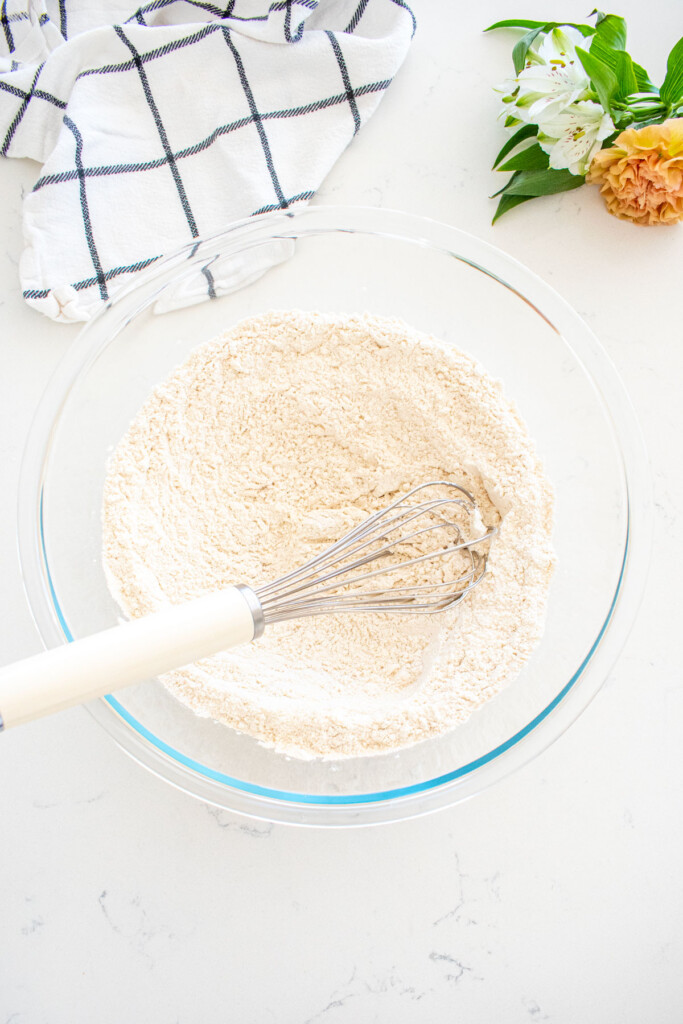 dry ingredients for buttermilk pumpkin waffles in a glass mixing bowl with a whisk on a marble counter with fresh flowers.
