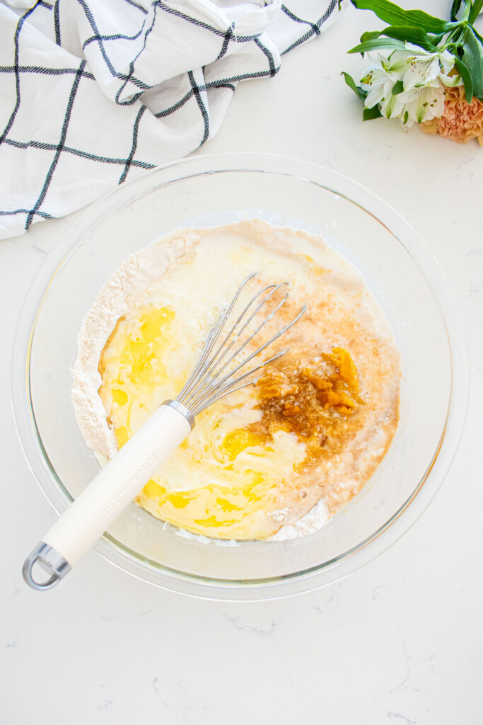 ingredients for buttermilk pumpkin waffles in a glass mixing bowl with a whisk on a marble counter with fresh flowers.