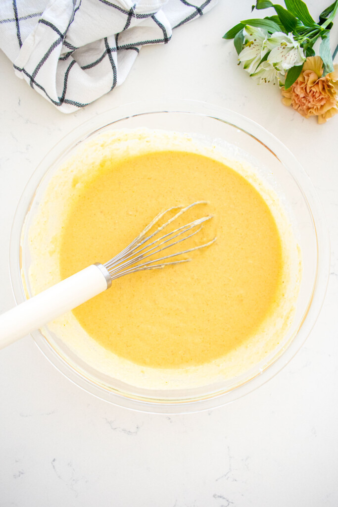 buttermilk pumpkin waffle batter in a glass mixing bowl with a whisk on a marble counter with fresh flowers.
