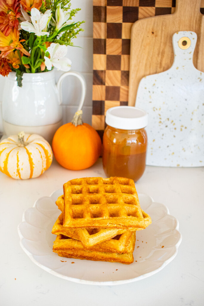 stack of buttermilk pumpkin waffles on a plate on a marble counter with small pumpkins and fresh flowers.