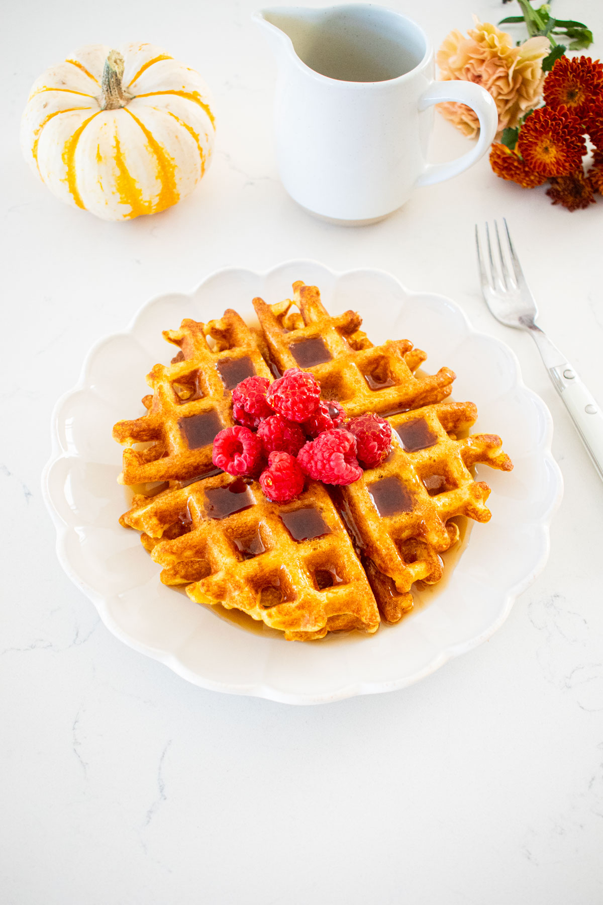 buttermilk pumpkin waffles on a plate with raspberries and maple syrup on a marble counter with fresh flowers.
