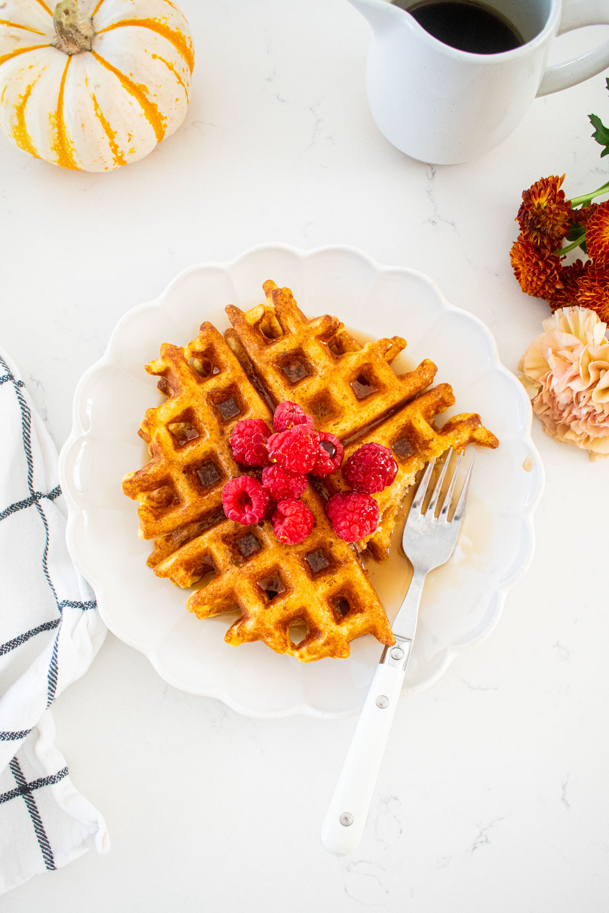 buttermilk pumpkin waffles on a plate with raspberries and maple syrup on a marble counter with fresh flowers.