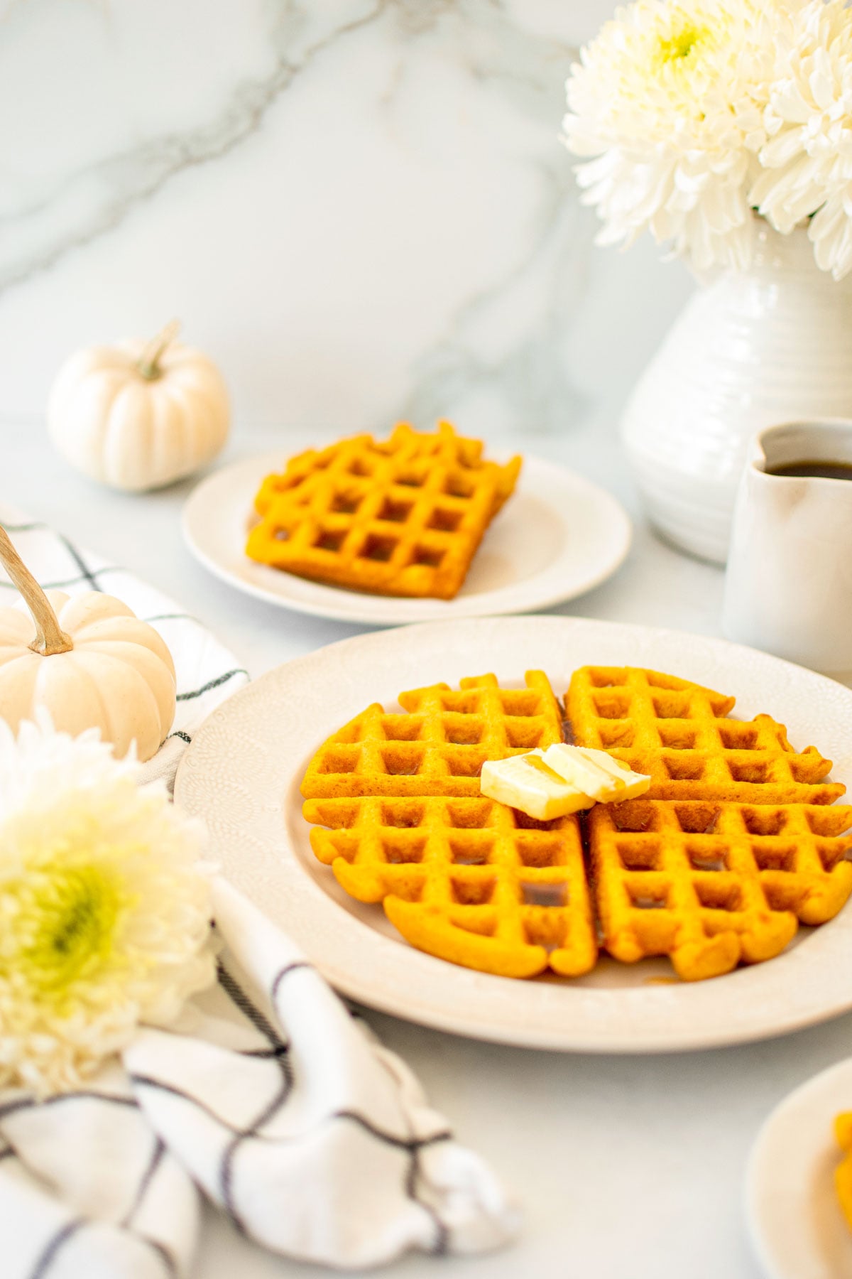 buttermilk pumpkin waffles on a white plate with butter on a white table with fresh flowers and maple syrup.