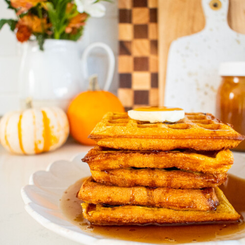 stack of buttermilk pumpkin waffles with butter and maple syrup on a marble counter with mini pumpkins and fresh flowers.