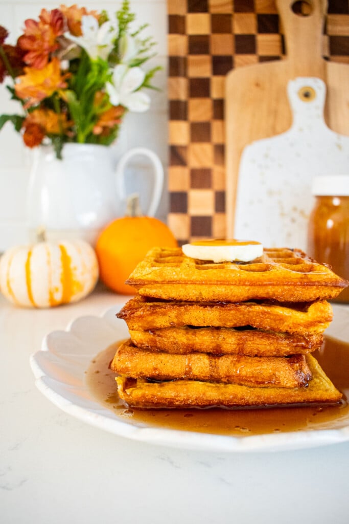 stack of buttermilk pumpkin waffles with butter and maple syrup on a marble counter with mini pumpkins and fresh flowers.
