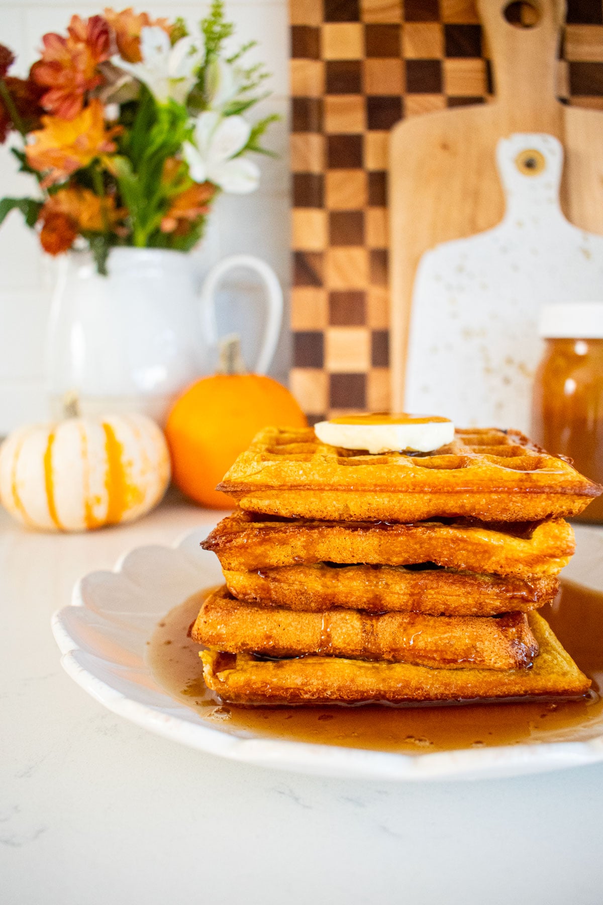 stack of buttermilk pumpkin waffles with butter and maple syrup on a marble counter with mini pumpkins and fresh flowers.