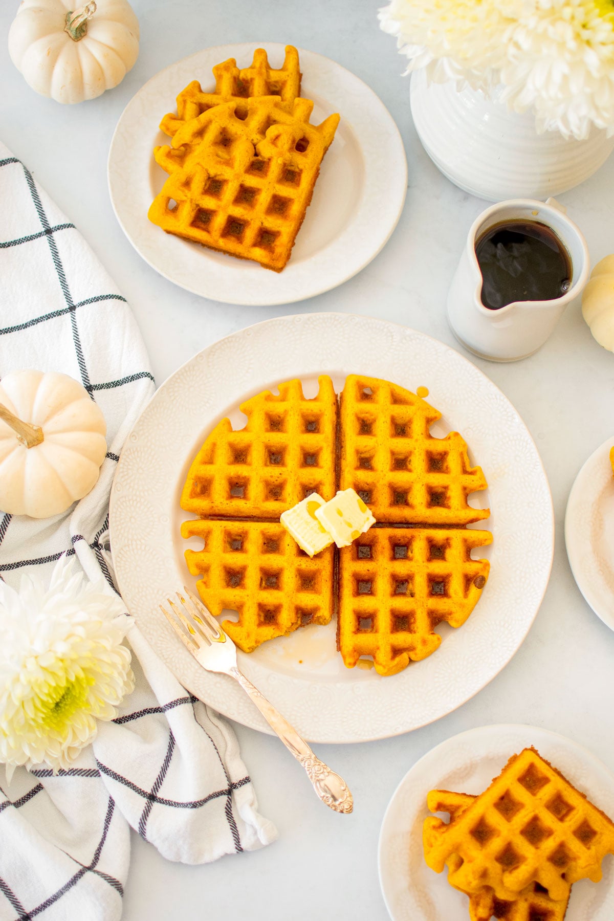 buttermilk pumpkin waffles on a white plate with butter on a white table with fresh flowers and maple syrup.