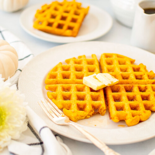 buttermilk pumpkin waffles on a white plate with butter on a white table with fresh flowers and maple syrup.