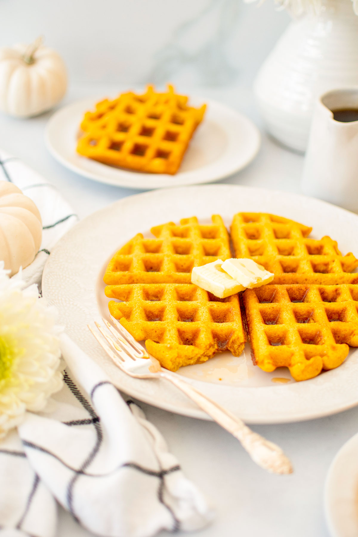buttermilk pumpkin waffles on a white plate with butter on a white table with fresh flowers and maple syrup.