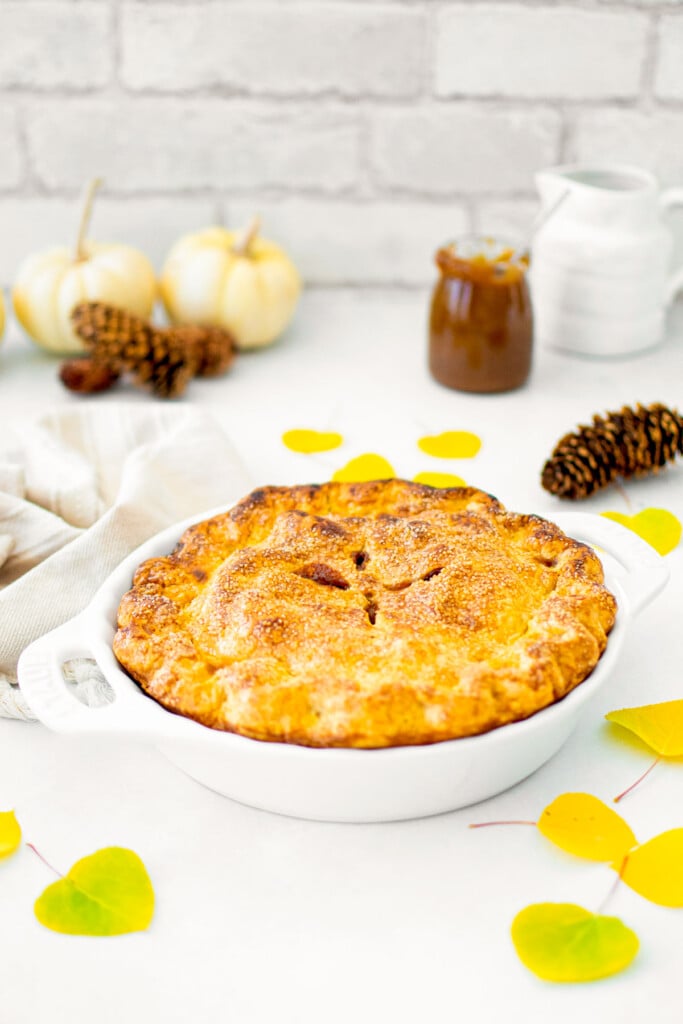 caramel apple pie on a white counter with fall leaves, pinecones and white pumpkins.