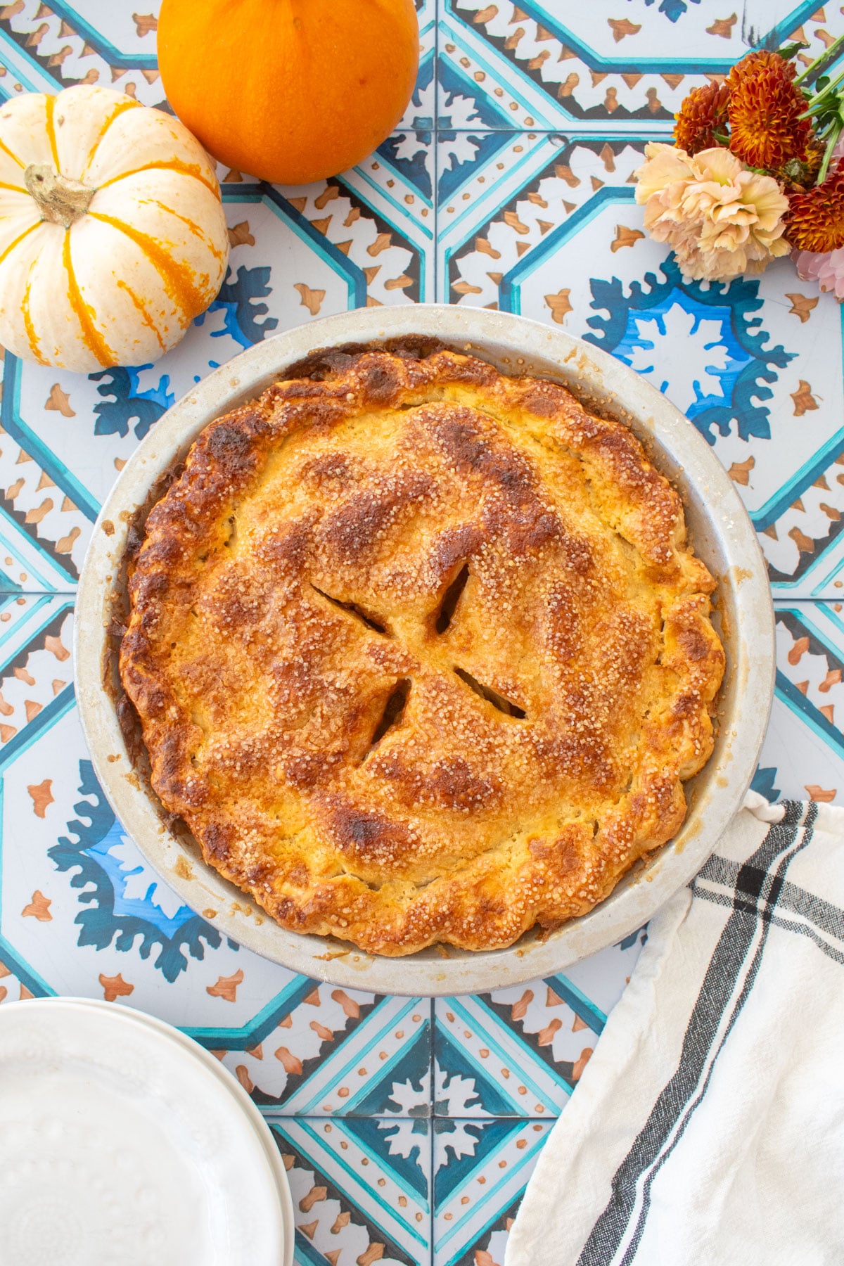 golden brown caramel apple pie on a tile table with mini pumpkins, fresh flowers and pie plates.