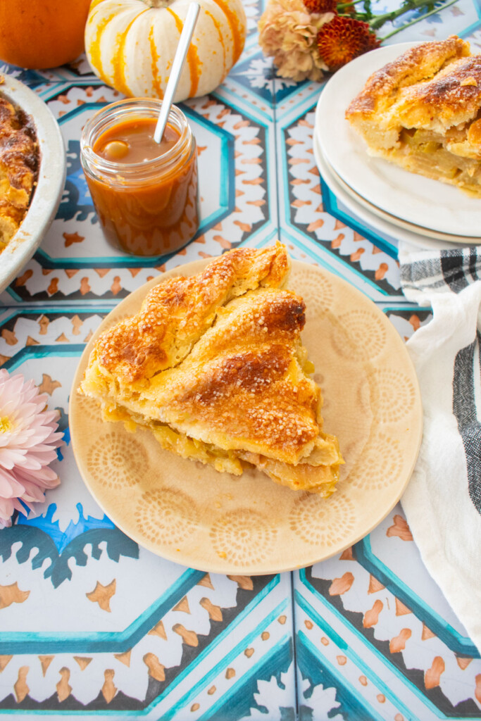 slice of caramel apple pie on a dessert plate with a jar of caramel, mini pumpkins and fresh flowers on a tile table.