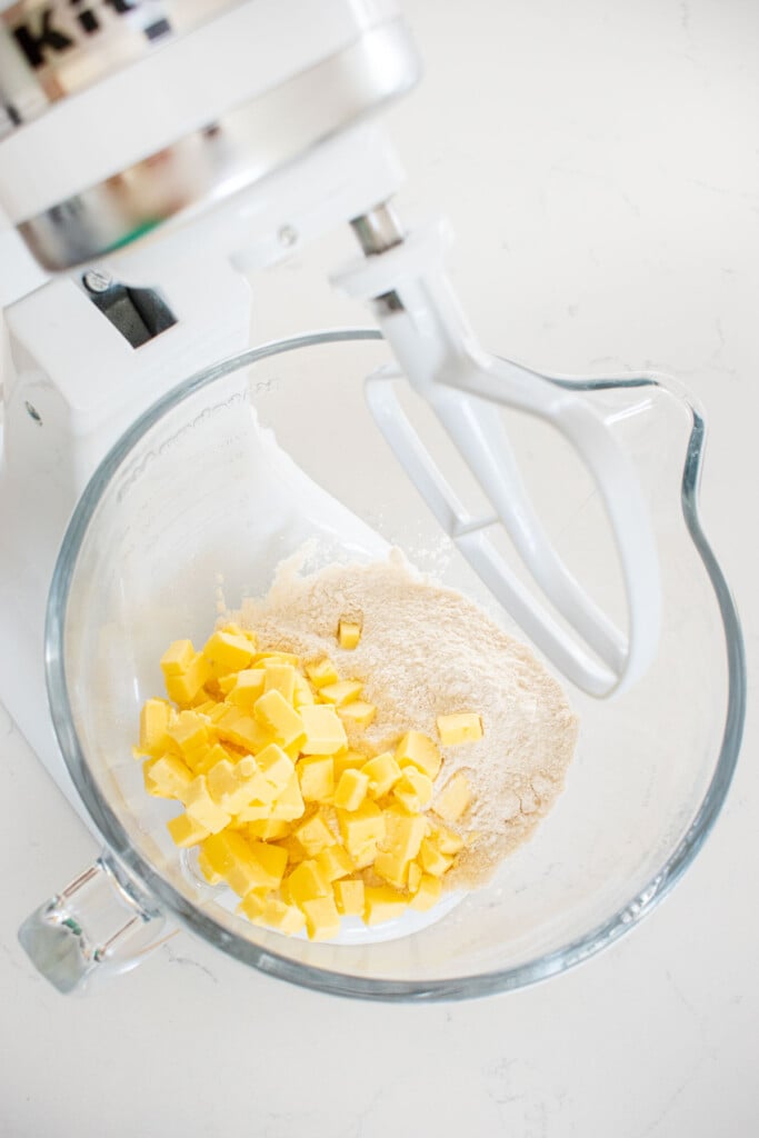 flour and cubed butter in a glass stand mixing bowl on a marble counter.