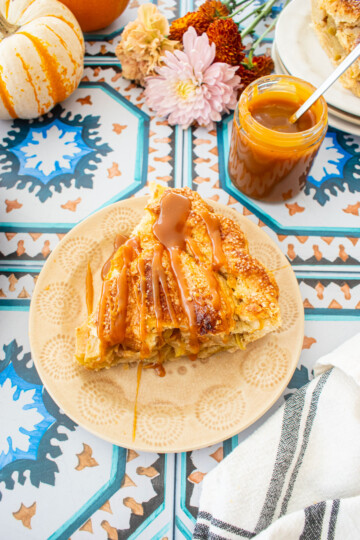 slice of caramel apple pie with a drizzle of caramel sauce on top on a tile table with mini pumpkins and fresh flowers.